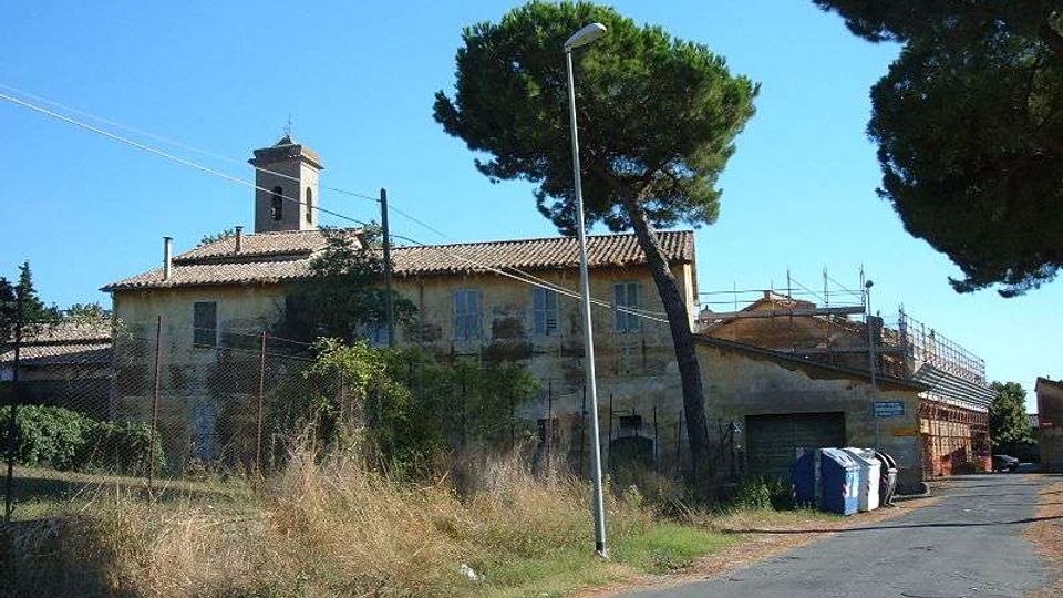 Edificio rurale a due piani con tetto in coppi e una piccola torre campanaria, circondato da recinzioni e vegetazione incolta. La scena è lungo una strada asfaltata con lampione, pini alti e alcuni cassonetti, sotto un cielo sereno.