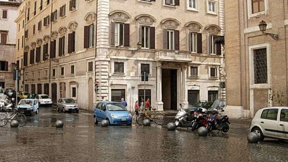 Facciata di un palazzo storico in pietra su una piazza lastricata in sampietrini, con finestre ad arco, persiane scure e un portale centrale. Davanti all’edificio si vedono auto e motocicli parcheggiati, dissuasori sferici e una strada urbana tra altri edifici in muratura.