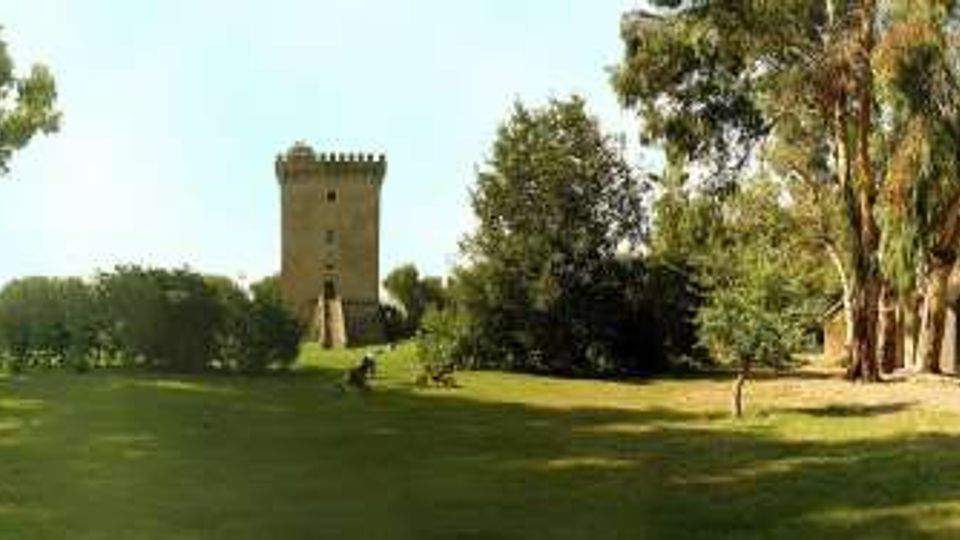 Ampio prato verde in un parco alberato, con una torre in pietra merlata al centro sullo sfondo. Ai lati si vedono un edificio in muratura rossastra e una struttura con archi, circondati da alberi alti.