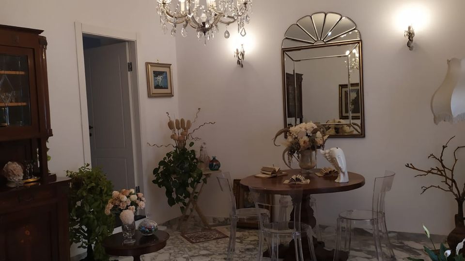 Sala da pranzo in stile classico con pavimento in marmo, grande lampadario di cristallo e pareti bianche con applique. Al centro c’è un tavolo rotondo con sedie trasparenti, specchio decorativo alla parete e mobili in legno con tappeti e piante ornamentali.