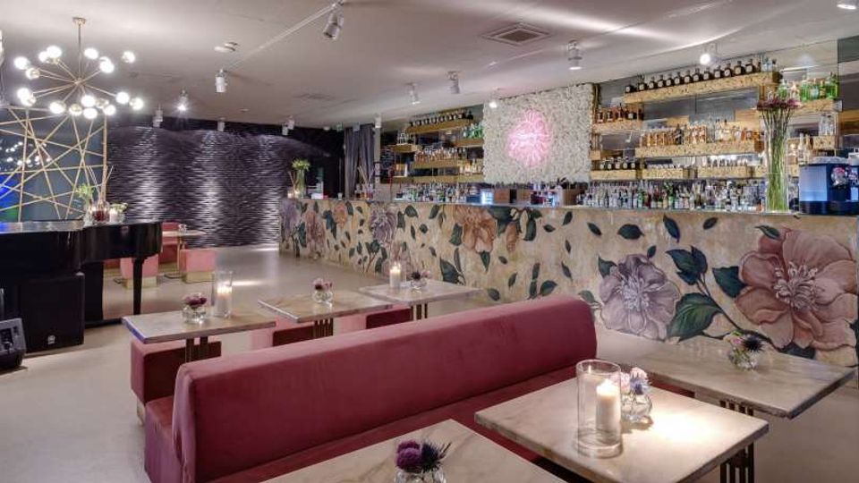 Interno di un bar-lounge moderno con divanetti rosa e tavolini in marmo illuminati da candele, e lungo bancone decorato con motivi floreali e scaffali di bottiglie sullo sfondo. A sinistra si vede un pianoforte a coda e lampadari sferici, con pareti scure a texture ondulata e illuminazione a faretti.
