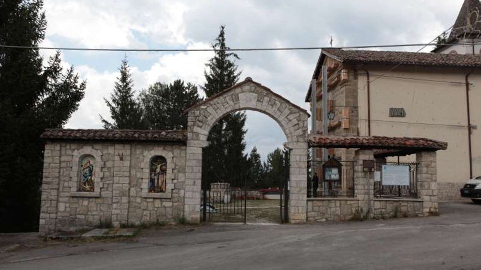 Ingresso in pietra di un complesso religioso con grande arco centrale e cancellata in ferro, affiancato da un muretto con nicchie decorate e da un edificio con tetto in coppi. Sullo sfondo si vedono alberi alti e cielo nuvoloso lungo una strada asfaltata.