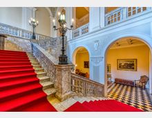 Interno di un edificio storico con ampia scalinata in marmo rivestita da tappeto rosso, balaustre in pietra e alti lampioni decorativi. Archi e pareti bianche con rilievi ornamentali si affacciano su un salone con pavimento a scacchi, quadri incorniciati e arredi classici.