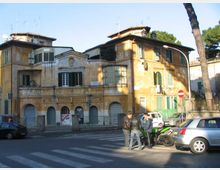Edificio storico a più piani con facciata ocra e persiane verdi, con portici ad archi al piano terra e finestre ad arco al primo piano. Davanti si vede un incrocio con strisce pedonali, auto parcheggiate e un lampione, con alberi sullo sfondo.