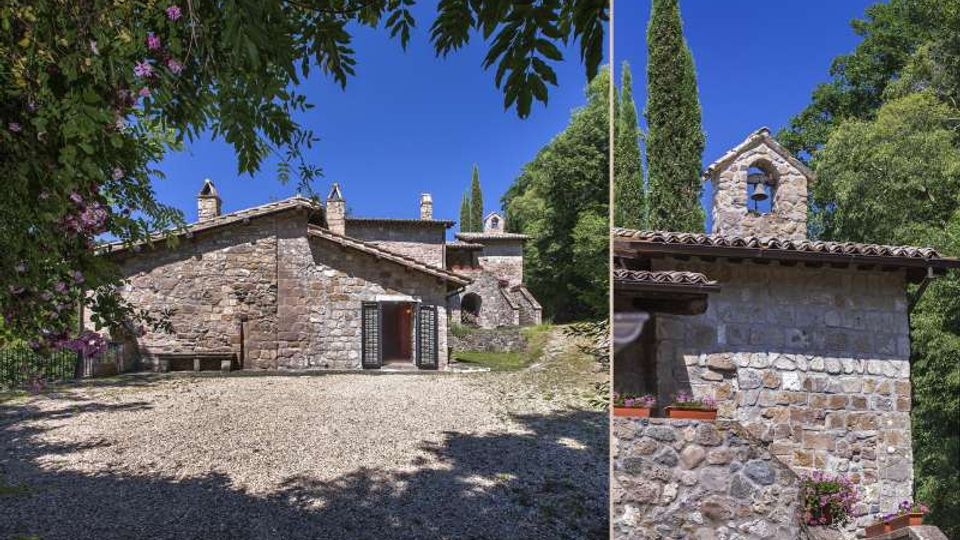 Complesso rurale in pietra con edifici a tetto in coppi e piccola struttura campanaria, affacciato su un cortile in ghiaia. Intorno si vedono alberi alti e vegetazione rigogliosa sotto un cielo sereno.