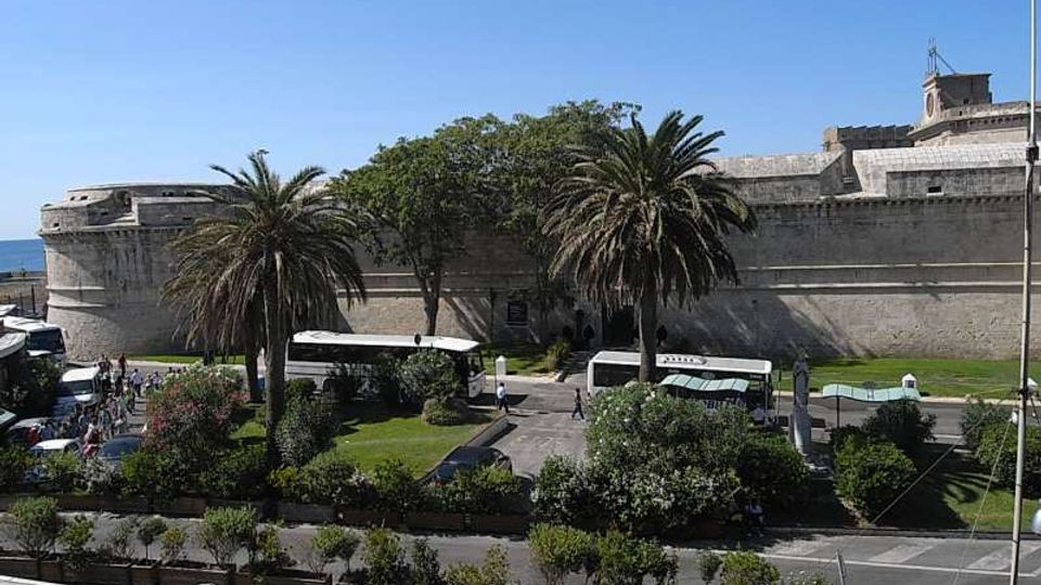 Veduta di una grande fortificazione in pietra con bastione arrotondato e mura alte, affacciata sul mare. In primo piano si vede un giardino urbano con palme, aiuole e vialetti, con una strada e alcuni autobus parcheggiati.