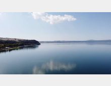 Ampio lago dalle acque calme con riflessi di nuvole, visto da lontano. Sulla sinistra si vede una costa boscosa con una linea di alberi, mentre all’orizzonte compaiono colline e una sponda distante sotto un cielo sereno.