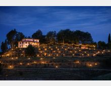 Casa di campagna su una collina al crepuscolo, circondata da alberi e da una fila di luci sospese. Il pendio è punteggiato da numerose lanterne accese disposte lungo i terrazzamenti.