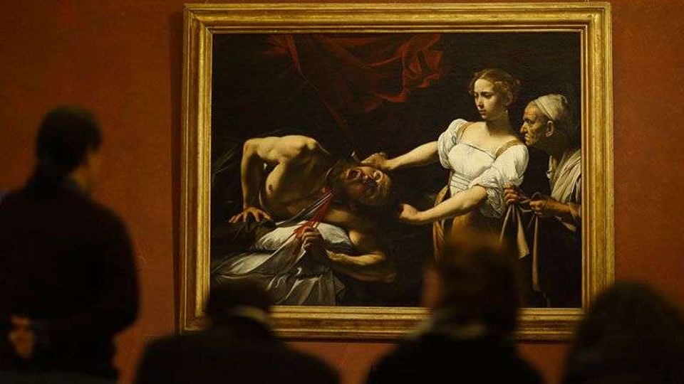 Sala museale con pareti color ocra e un grande dipinto incorniciato in oro appeso al centro. Davanti al quadro si intravede il profilo sfocato di visitatori, mentre l’opera raffigura una scena drammatica con figure illuminate su sfondo scuro.