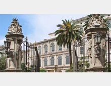 Ingresso monumentale con cancelli in ferro battuto tra due pilastri in pietra decorati con sculture e lanterne. Sullo sfondo si vede un grande edificio storico a più piani con finestre ad arco e palme nel cortile.