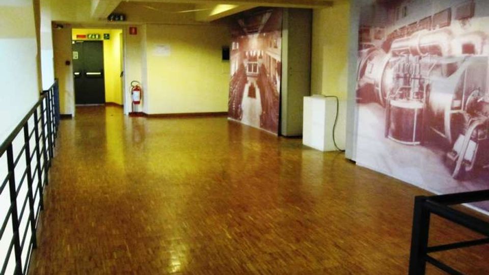 Ampio corridoio interno con pavimento in parquet lucido, pareti gialle e ringhiera metallica nera sul lato sinistro. Sul fondo si vede una porta verde con segnaletica di uscita e un estintore, mentre sulla parete destra sono presenti grandi pannelli fotografici.