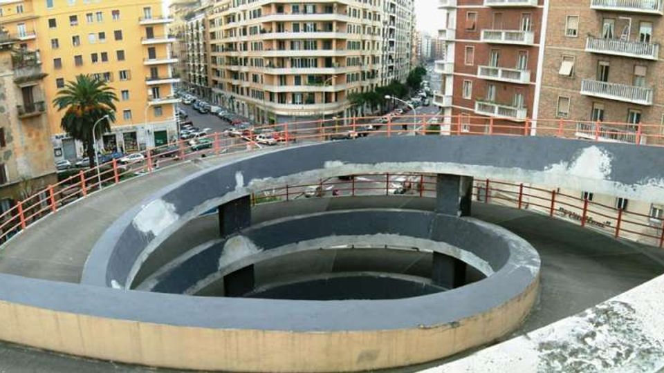 Rampa pedonale sopraelevata a spirale in cemento, con parapetti metallici, vista dall’alto. Sullo sfondo si vedono strade urbane e palazzi residenziali multipiano con balconi.