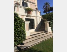Facciata di una villa in stile classico con pareti rosa e bianche, balaustra e grandi vasi di terracotta. In primo piano una scalinata in pietra conduce a un ingresso con cancellata in ferro battuto, affiancata da siepi e palme in giardino.