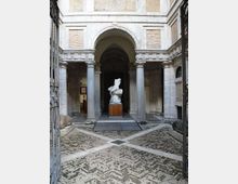 Cortile interno di un edificio storico con pavimentazione in pietra a motivi geometrici, colonne in pietra e un grande arco centrale che introduce a un portico voltato. Al centro è esposta una statua su basamento, circondata da pareti in laterizio e intonaco con aperture laterali.