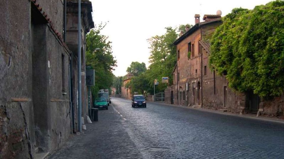 Strada urbana in pavé con marciapiede stretto, fiancheggiata da edifici in muratura dall’aspetto storico. Sul lato destro si vede un edificio con finestre e alberi che coprono parte della facciata, con alcune auto lungo la carreggiata.