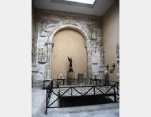 Sala museale con grande arco in pietra scolpita e iscrizioni, incassato in una parete color ocra e illuminato da un lucernario. Davanti all’arco sono esposti reperti e statue su piedistalli, con una struttura metallica che delimita una grande base circolare al centro.