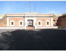 Facciata simmetrica in mattoni rossi con cornici in pietra bianca e ingresso ad arco centrale con cancello, sormontato da un’insegna e da due aste portabandiera. Davanti si estende un ampio piazzale asfaltato, con mura laterali che incorniciano l’accesso.