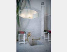 Interno di un bagno in marmo con vasca incassata e rubinetteria dorata, affiancata da due piccoli sgabelli con cuscini. Sulla parete c’è uno specchio esagonale con luci riflesse e una colonna in marmo vicino al bordo della vasca.