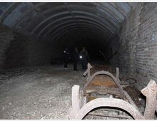 Interno di una galleria sotterranea in mattoni con soffitto a volta, pavimento in ghiaia e pareti laterali scure. In primo piano si vedono strutture metalliche arrugginite e detriti, mentre il tunnel si estende in profondità verso un’area poco illuminata.