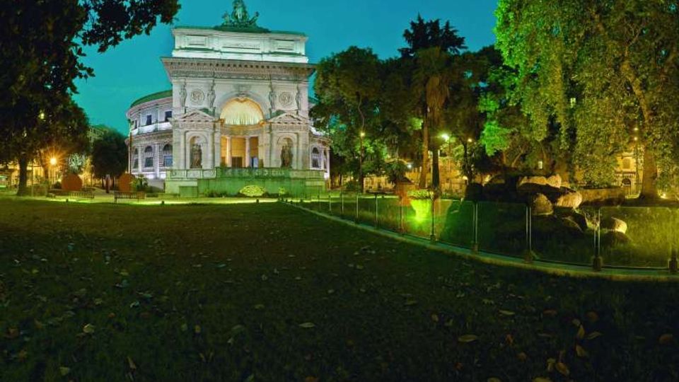 Edificio monumentale in stile classico illuminato di notte, con grande arco centrale e colonne, circondato da un parco alberato. In primo piano prato e percorso con illuminazione verde e recinzione bassa, con massi e vegetazione sul lato destro.