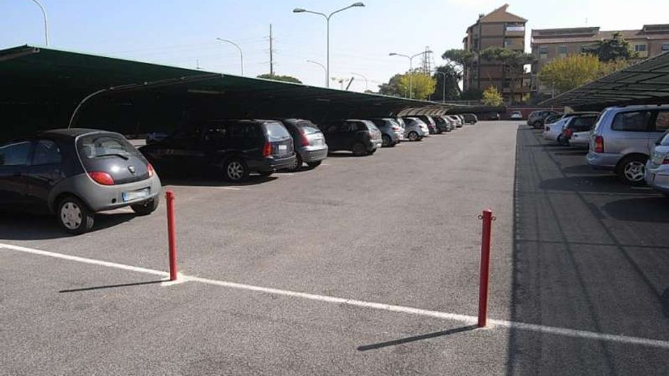Parcheggio esterno asfaltato con file di auto sotto pensiline metalliche su entrambi i lati e corsia centrale ampia. Sullo sfondo si vedono lampioni stradali e alcuni edifici tra alberi.