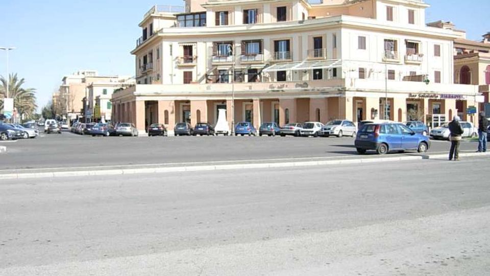Ampia piazza asfaltata con parcheggio e auto davanti a un grande edificio d’angolo a più piani, con facciata chiara, balconi e portici al piano terra con negozi. Sullo sfondo si vedono altri palazzi lungo la strada e alcune palme ai margini.