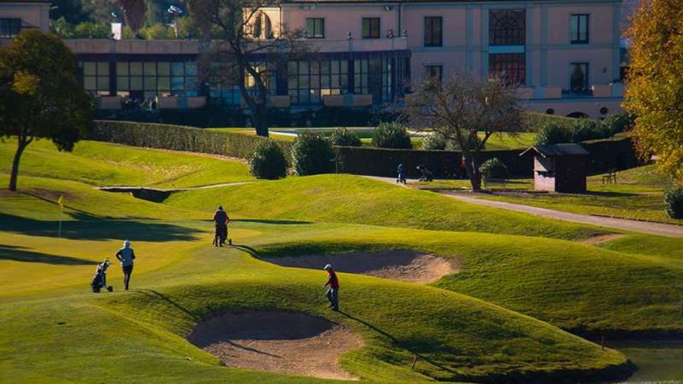Campo da golf con colline erbose ondulate e bunker di sabbia in primo piano, attraversato da un vialetto. Sullo sfondo si vede un grande edificio in stile villa con facciata chiara e ampie finestre, circondato da alberi e siepi.