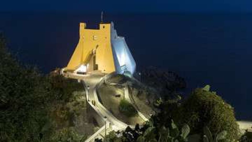 Edificio bianco dalle forme moderne e angolari, illuminato di notte, su un promontorio roccioso affacciato sul mare. Una strada tortuosa e una scalinata conducono all’ingresso, circondate da vegetazione mediterranea.