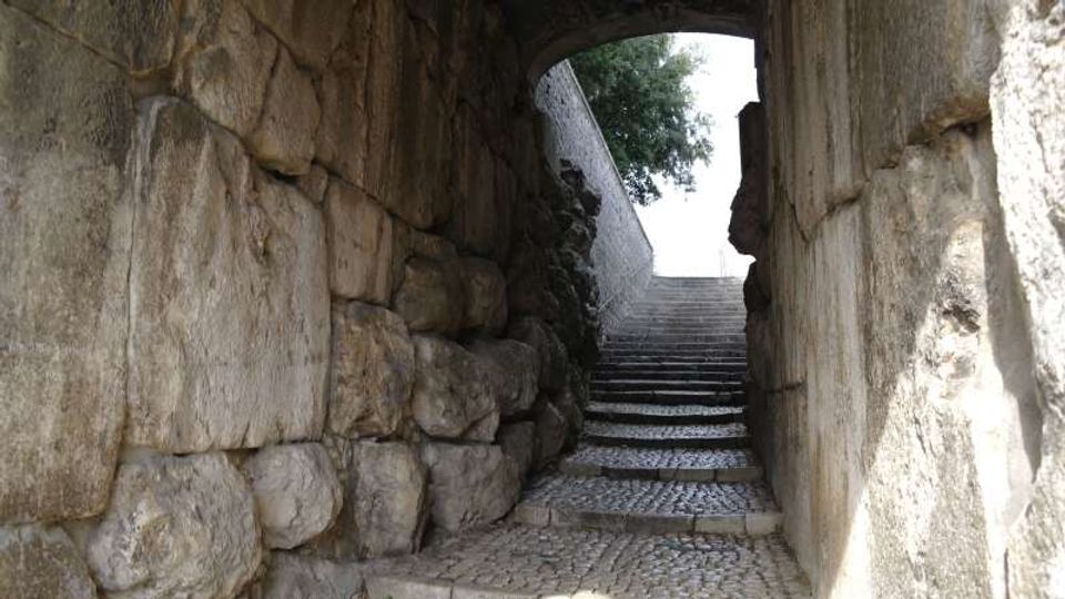 Passaggio in pietra con pareti di grandi blocchi e soffitto ad arco, che conduce a una scalinata in ciottoli e gradini in pietra. In cima si apre un’uscita luminosa verso l’esterno, con un albero visibile oltre l’arco.
