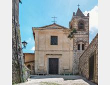 Facciata di una piccola chiesa in pietra con portale centrale, finestra quadrata e due croci sul tetto, affiancata da un campanile con aperture ad arco e cuspide con croce. Davanti c’è una piazzetta lastricata in ciottoli, circondata da muri in pietra e una lanterna a parete sotto cielo azzurro.