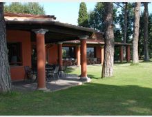Veranda coperta di una struttura in stile rustico con colonne cilindriche color terracotta e tetto in tegole, arredata con tavoli e sedie. L’edificio è circondato da un prato verde con alti pini e alberi sullo sfondo.