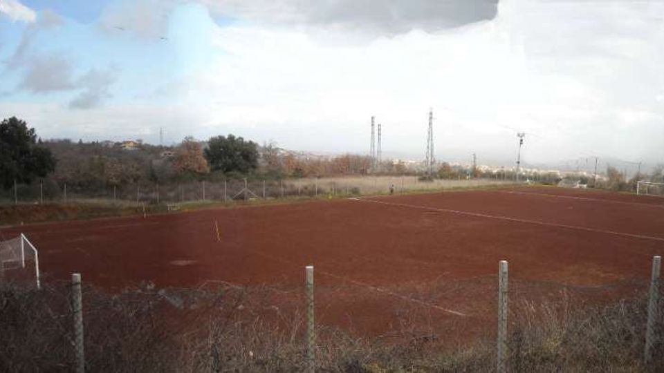 Campo da calcio in terra rossa recintato, con porte bianche e linee del terreno visibili. Sullo sfondo si vedono alberi, tralicci e un paesaggio rurale sotto un cielo nuvoloso.