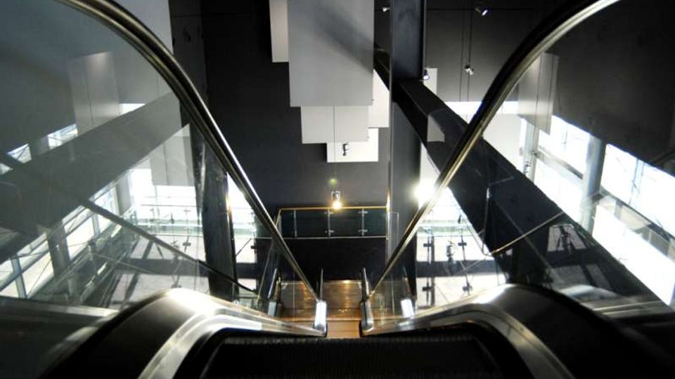 Vista dall’alto di una scala mobile in un atrio moderno con pareti scure e ampie superfici vetrate. Si vedono ringhiere metalliche lucide, finestre laterali che lasciano entrare luce naturale e pannelli geometrici sospesi sul soffitto.