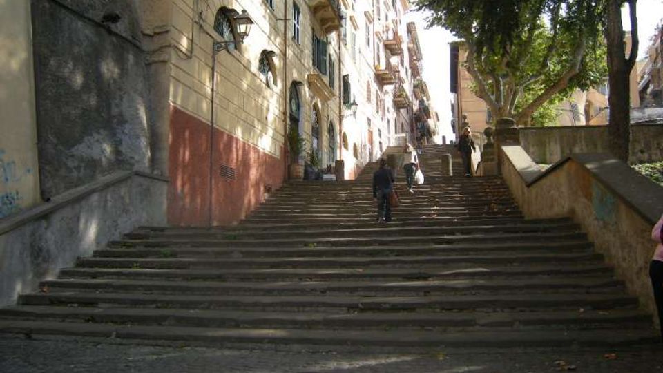 Ampia scalinata in pietra in un contesto urbano, affiancata da palazzi residenziali con persiane e piccoli balconi sul lato sinistro. Sul lato destro ci sono alberi e un muretto che costeggia i gradini, con luce solare che filtra tra le fronde.