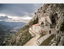 Santuario in pietra addossato a una parete rocciosa su un ripido versante montano, con terrazze murate, arco d’ingresso e sentiero curvo che costeggia l’edificio. Sullo sfondo si vede una valle ampia con colline e un cielo nuvoloso.