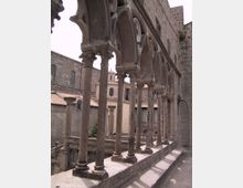 Loggiato in pietra di un edificio storico, con una fila di colonne sottili e archi decorati che affacciano su un cortile interno. Sullo sfondo si vedono facciate in muratura e tetti in tegole di altri edifici del complesso.