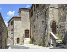 Vicolo in pietra in un borgo storico, con edifici in muratura grezza e facciate alte affacciate su una piccola piazzetta. Si vedono porte ad arco, finestre strette, una scalinata con corrimano in pietra, lampioni a parete e pavimentazione in lastre sotto un cielo azzurro.