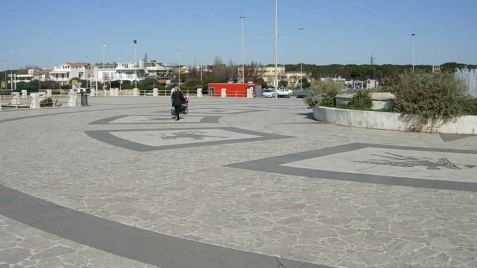 Ampia piazza pavimentata in pietra con grandi fasce geometriche scure e motivi decorativi incisi sul suolo, illuminata da alti lampioni. Sullo sfondo si vedono aiuole con vegetazione bassa, parcheggi e edifici bassi sotto un cielo sereno.