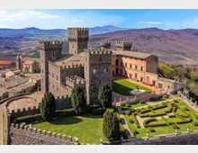 Veduta aerea di un castello medievale in pietra con torri merlate e mura fortificate, affiancato da un edificio in intonaco color ocra. Nel cortile interno e nei giardini geometrici con siepi potate si vede una fontana, con un borgo e colline montuose sullo sfondo.
