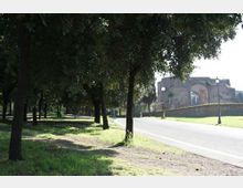 Viale alberato con ampie aree erbose e una strada asfaltata che curva verso destra. Sullo sfondo si vede una struttura storica in muratura con grandi archi, oltre un muretto e alcuni lampioni.