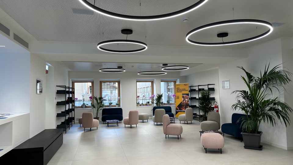 Ampia sala d’attesa moderna con pavimento chiaro, pareti bianche e grandi lampade circolari a LED sospese. Sono presenti poltroncine e divanetti dai colori neutri, scaffalature laterali, piante in vaso e finestre sul fondo che illuminano l’ambiente.