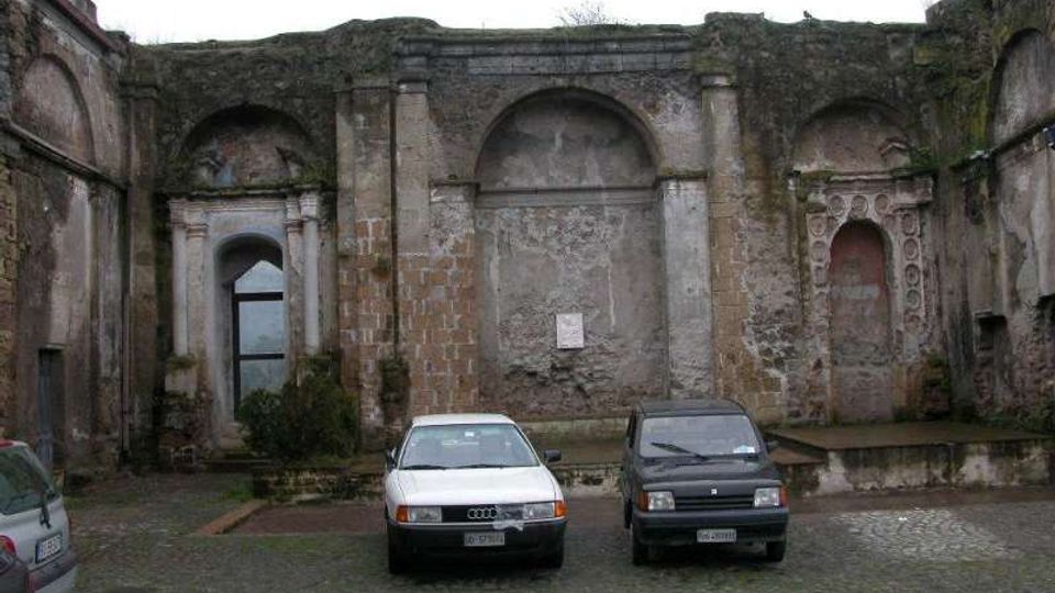 Cortile lastricato in sampietrini con una facciata in muratura antica e deteriorata, scandita da grandi nicchie ad arco e pilastri in pietra. Sullo sfondo si notano un portale ad arco con infisso vetrato e parti di pareti scrostate con tracce decorative.