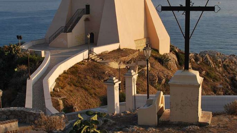 Antica torre costiera intonacata di bianco su un promontorio roccioso affacciato sul mare, raggiunta da una rampa e una scalinata con parapetti. In primo piano si vede una grande croce nera su piedistallo e alcuni muretti e lampioni lungo il percorso.