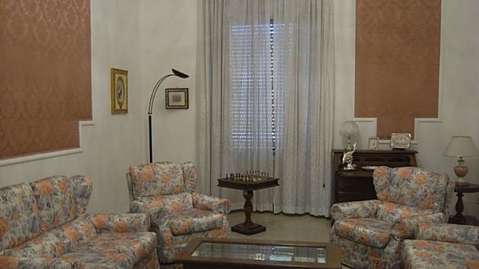 Salotto interno con divani e poltrone imbottiti a fantasia floreale disposti attorno a un tavolino rettangolare con piano in vetro su tappeto decorato. Sulla parete di fondo c’è una grande portafinestra con tende leggere, con lampade e mobili d’appoggio ai lati e pannelli decorativi alle pareti.