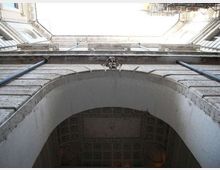 Veduta dal basso di un cortile interno tra edifici in muratura chiara, con grande arco d’ingresso in primo piano e cornici in pietra. Sotto l’arco si intravede un soffitto decorato a cassettoni e lungo le pareti corrono tubazioni verticali.