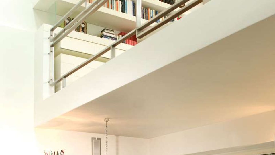 Soggiorno moderno a doppia altezza con soppalco e ringhiera metallica, pareti bianche e grandi scaffali pieni di libri. In basso si vede una poltrona sospesa trasparente e una grande finestra con vista su un piccolo giardino e un muro in mattoni.