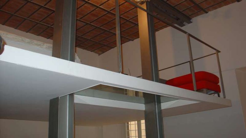 Interno di un loft su due livelli con soppalco in metallo e pavimento bianco, sostenuto da pilastri scuri e protetto da ringhiera. Soffitto alto con travi e mattoni a vista, illuminazione a spot e finestra verticale con vetri a riquadri.