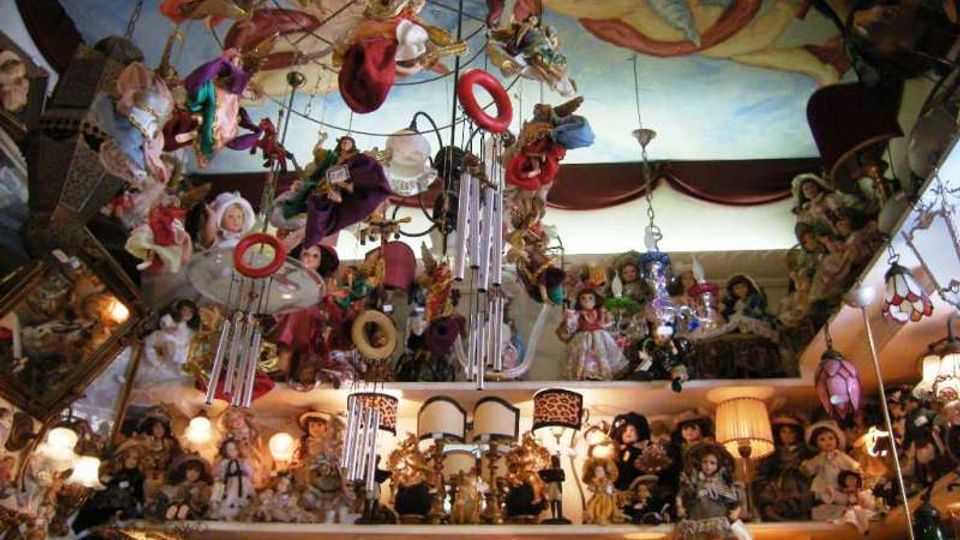 Interno di un negozio o stanza collezionistica molto affollata, con scaffali pieni di bambole e numerose lampade decorative accese. Dal soffitto dipinto con un affresco pendono oggetti sospesi come campanelli eolici, cerchi e decorazioni.