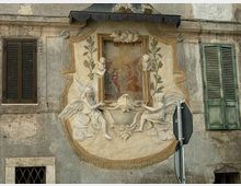 Facciata di un edificio storico con un riquadro decorativo in stucco che incornicia un piccolo affresco centrale, affiancato da figure angeliche e motivi vegetali in rilievo. Ai lati si vedono due finestre con persiane in legno e un cartello stradale in primo piano.