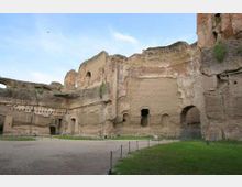 Ampia area archeologica con grandi rovine in mattoni e pareti scavate, con arcate e aperture nella muratura. In primo piano si vedono un sentiero e una zona erbosa delimitata da paletti e corde sotto un cielo aperto.